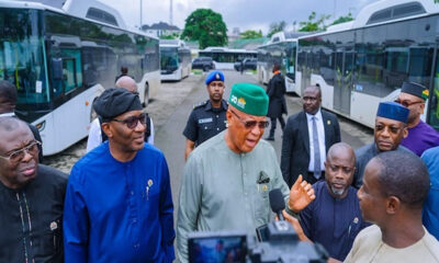 Group of Nigerian officials, including a man in a green cap, addressing reporters beside waiting buses at a transport yard.