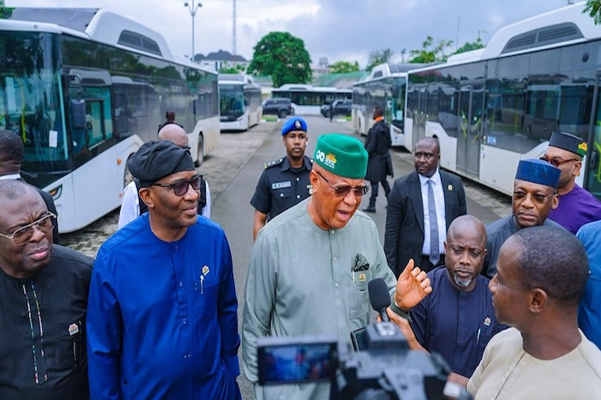 Group of Nigerian officials, including a man in a green cap, addressing reporters beside waiting buses at a transport yard.