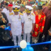 Group of officials at a ribbon-cutting ceremony, two men in white naval uniforms center, blue balloons in foreground.