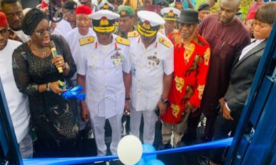Group of officials at a ribbon-cutting ceremony, two men in white naval uniforms center, blue balloons in foreground.