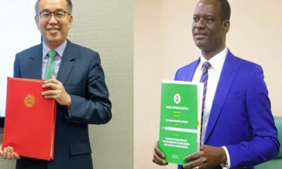 Two men in suits stand side by side, each holding a large colorful folder (red on the left, green on the right).