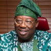 Smiling man in green traditional attire and glasses, seated in a red chair.