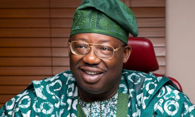 Smiling man in green traditional attire and glasses, seated in a red chair.
