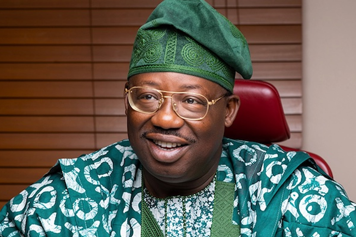 Smiling man in green traditional attire and glasses, seated in a red chair.