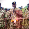 Three camouflaged soldiers at an outdoor ribbon-cutting ceremony, central officer speaking into a mic as the pink ribbon is cut by others nearby.