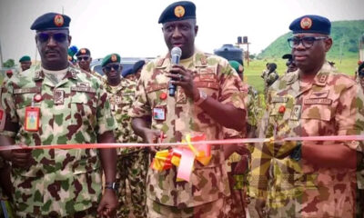 Three camouflaged soldiers at an outdoor ribbon-cutting ceremony, central officer speaking into a mic as the pink ribbon is cut by others nearby.