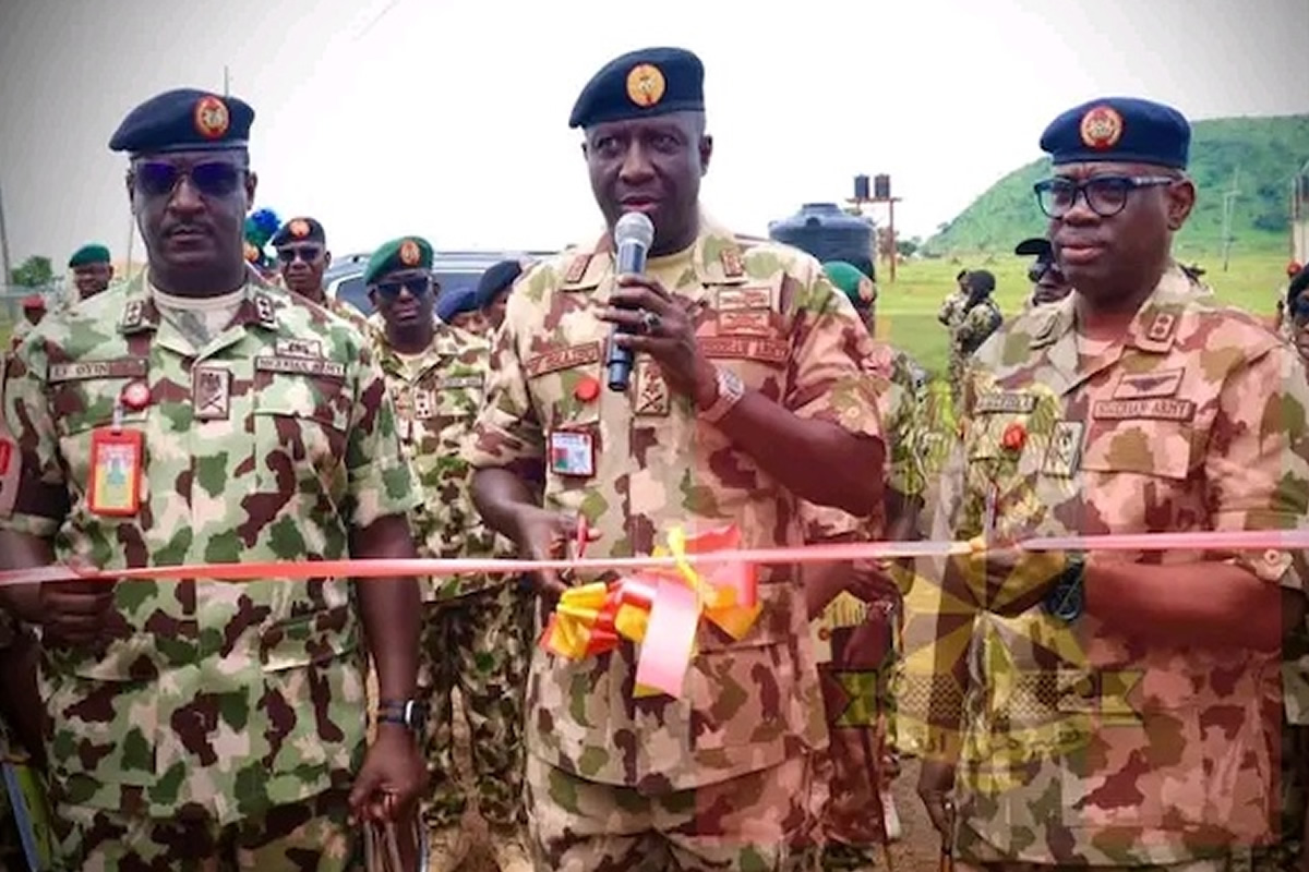 Three camouflaged soldiers at an outdoor ribbon-cutting ceremony, central officer speaking into a mic as the pink ribbon is cut by others nearby.