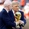 Two men in dark suits hold a large gold FIFA World Cup trophy during a ceremony with a blurred crowd in the background.