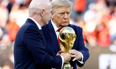Two men in dark suits hold a large gold FIFA World Cup trophy during a ceremony with a blurred crowd in the background.