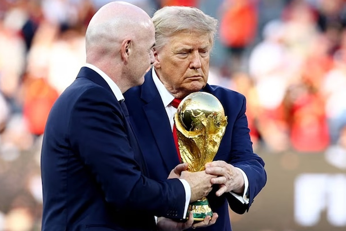 Two men in dark suits hold a large gold FIFA World Cup trophy during a ceremony with a blurred crowd in the background.