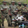 Group of camouflaged soldiers outdoors, a kneeling member holds a white quadcopter drone while others look on.