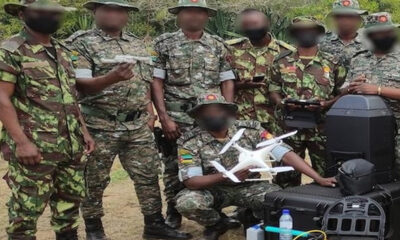 Group of camouflaged soldiers outdoors, a kneeling member holds a white quadcopter drone while others look on.