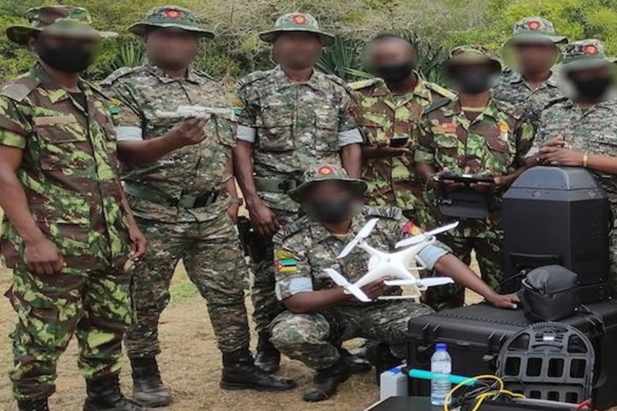 Group of camouflaged soldiers outdoors, a kneeling member holds a white quadcopter drone while others look on.