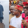 Two-panel scene: a tall statue in a blue robe on a pedestal left, and a man in a red cap speaking into a pink microphone at a crowd on the right.