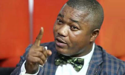 Man in a blue plaid suit with a green bow tie points a finger, speaking emphatically.