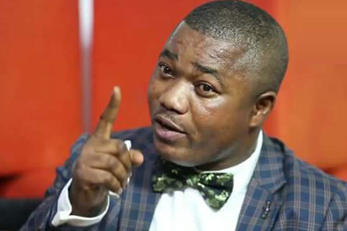 Man in a blue plaid suit with a green bow tie points a finger, speaking emphatically.