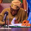 Elderly man in traditional attire signs documents at a desk with a microphone nearby, assisted by a standing person in a blue shirt.