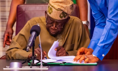 Elderly man in traditional attire signs documents at a desk with a microphone nearby, assisted by a standing person in a blue shirt.
