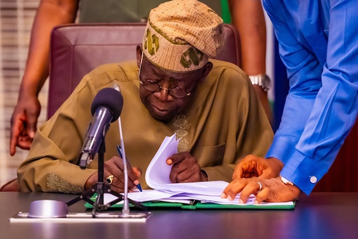 Elderly man in traditional attire signs documents at a desk with a microphone nearby, assisted by a standing person in a blue shirt.