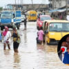 People wading in knee-deep floodwater on a flooded street with submerged cars; circular inset shows a man in a suit speaking at a podium.
