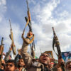 Crowd of protesters raising rifles overhead during a demonstration, with flags and framed photos visible in the background under a blue sky.