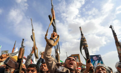 Crowd of protesters raising rifles overhead during a demonstration, with flags and framed photos visible in the background under a blue sky.