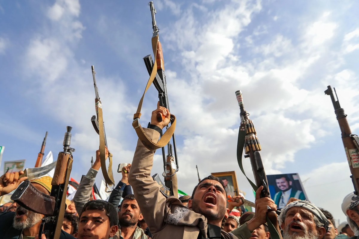 Crowd of protesters raising rifles overhead during a demonstration, with flags and framed photos visible in the background under a blue sky.