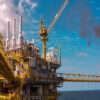 Offshore oil platform with a gas flare burning high, yellow scaffolding and a crane against a blue sky over the sea.