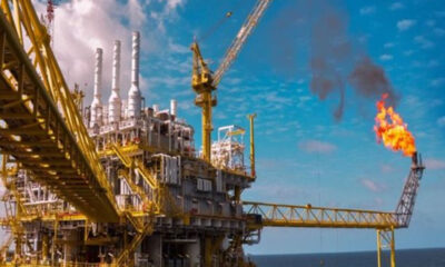 Offshore oil platform with a gas flare burning high, yellow scaffolding and a crane against a blue sky over the sea.