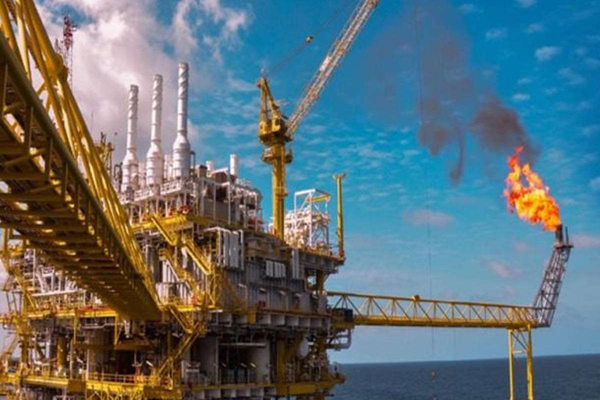 Offshore oil platform with a gas flare burning high, yellow scaffolding and a crane against a blue sky over the sea.