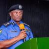 Police officer in blue uniform speaks at a green podium, holding an orange microphone.
