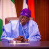 Older man in blue traditional attire sits at a desk with a microphone, flags in the background in a formal setting.