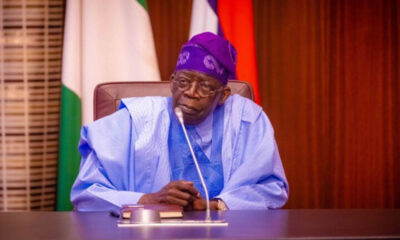 Older man in blue traditional attire sits at a desk with a microphone, flags in the background in a formal setting.