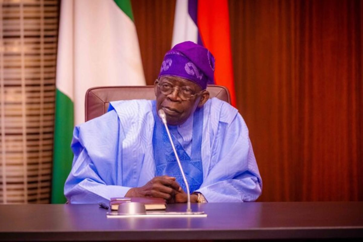 Older man in blue traditional attire sits at a desk with a microphone, flags in the background in a formal setting.