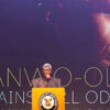 Man delivering a speech at a yellow podium with a crest, in front of a backdrop that reads 'Sanwo-Olu Against All Odds'