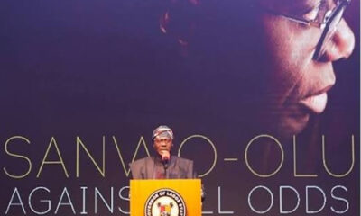 Man delivering a speech at a yellow podium with a crest, in front of a backdrop that reads 'Sanwo-Olu Against All Odds'