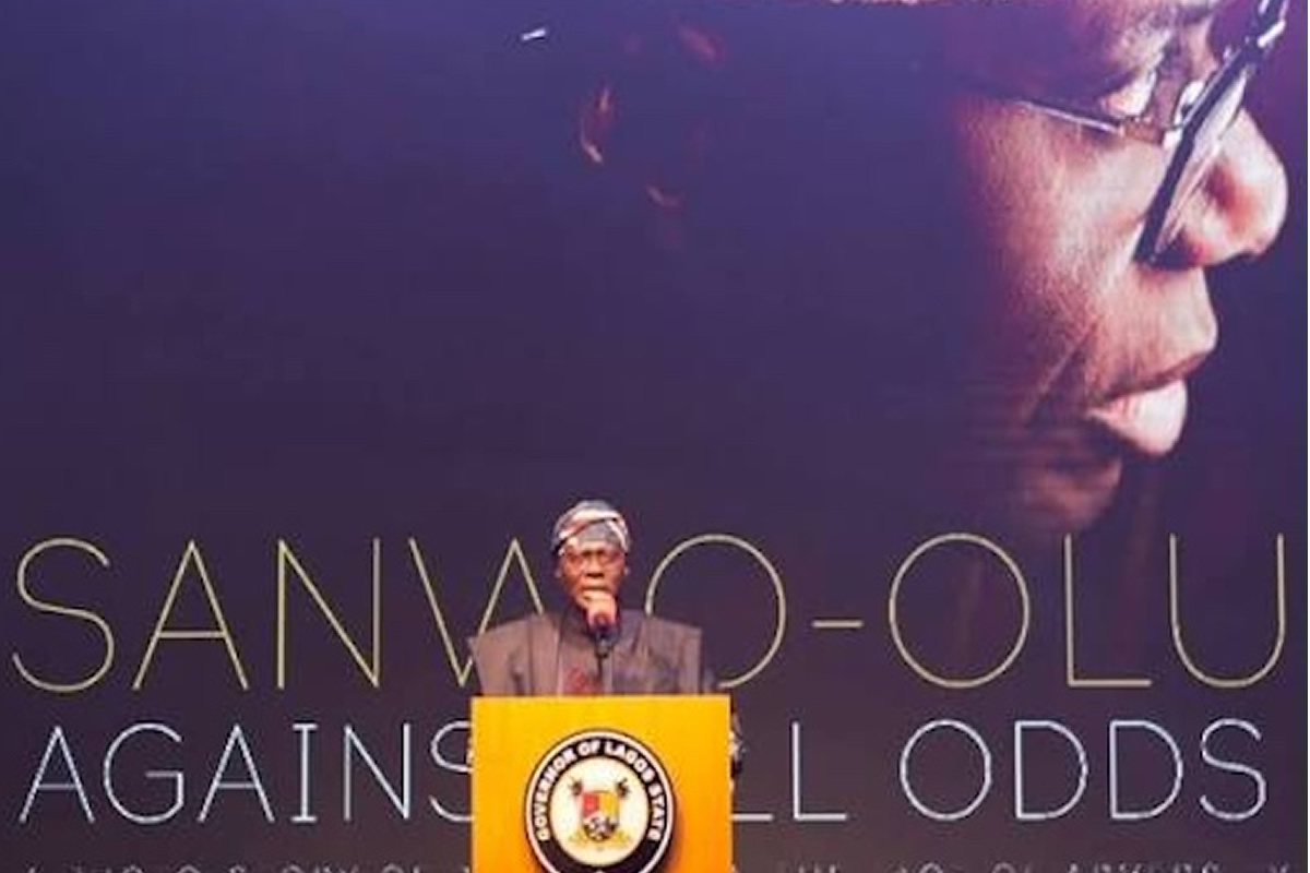 Man delivering a speech at a yellow podium with a crest, in front of a backdrop that reads 'Sanwo-Olu Against All Odds'