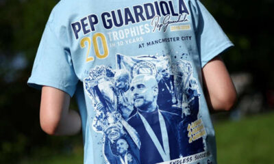 Back view of a person wearing a light-blue T-shirt celebrating Pep Guardiola's 20 trophies with trophy images on the shirt logo.