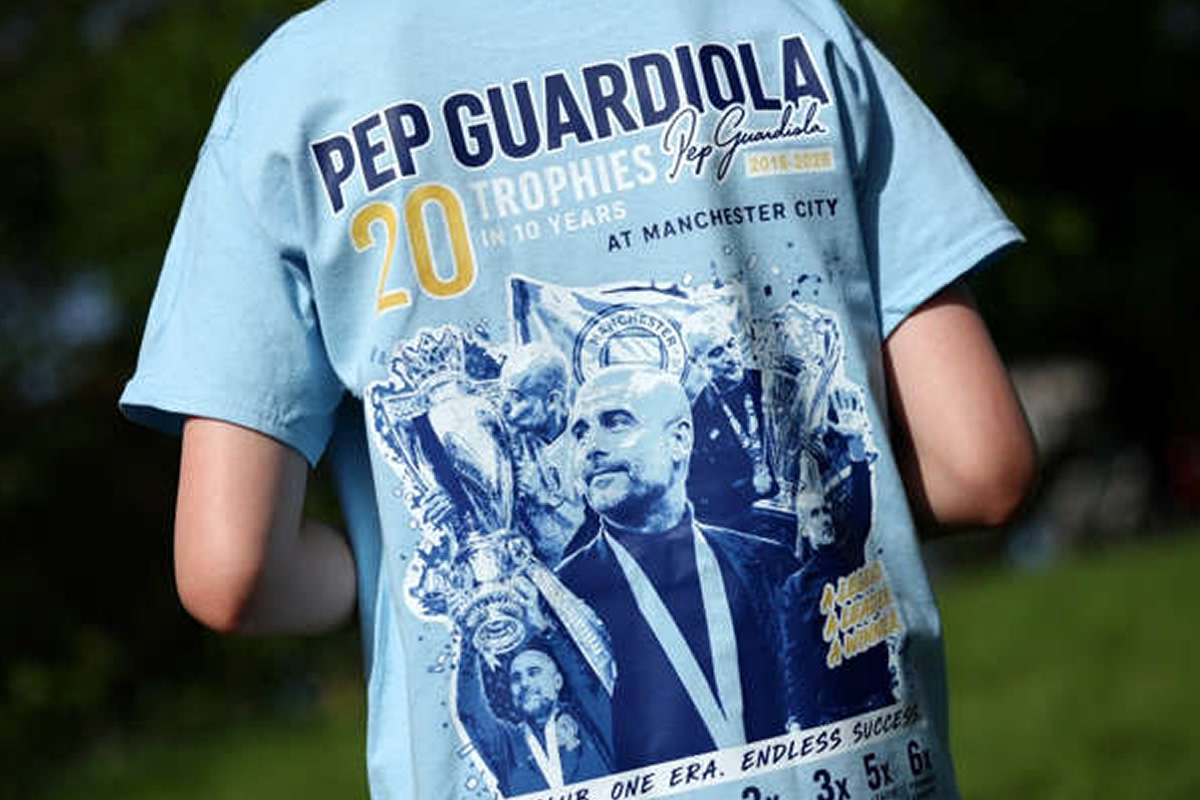 Back view of a person wearing a light-blue T-shirt celebrating Pep Guardiola's 20 trophies with trophy images on the shirt logo.