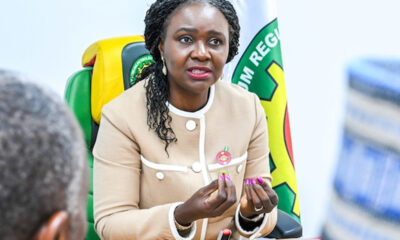 Woman in a beige blazer speaks at a meeting, hands gesturing as she talks, with a colorful flag in the background.