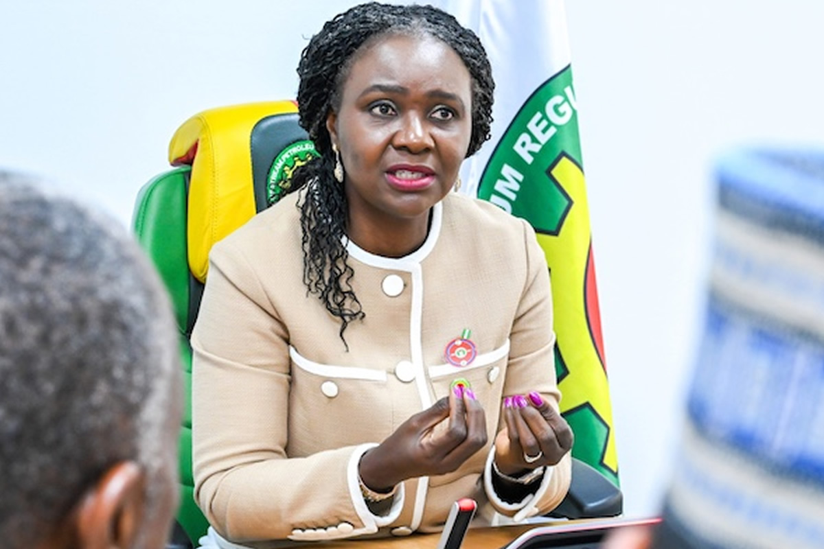Woman in a beige blazer speaks at a meeting, hands gesturing as she talks, with a colorful flag in the background.