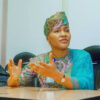 Woman in a colorful traditional outfit and headwrap sits at a conference table, gesturing with her hands as she speaks in a meeting.