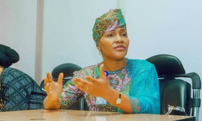 Woman in a colorful traditional outfit and headwrap sits at a conference table, gesturing with her hands as she speaks in a meeting.