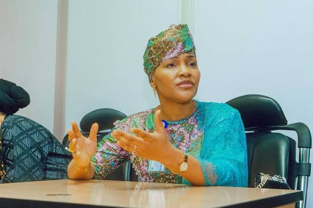 Woman in a colorful traditional outfit and headwrap sits at a conference table, gesturing with her hands as she speaks in a meeting.