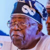 Elder man wearing a patterned head wrap and glasses, speaking into a microphone at an event.