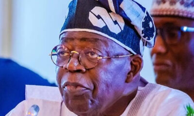Elder man wearing a patterned head wrap and glasses, speaking into a microphone at an event.