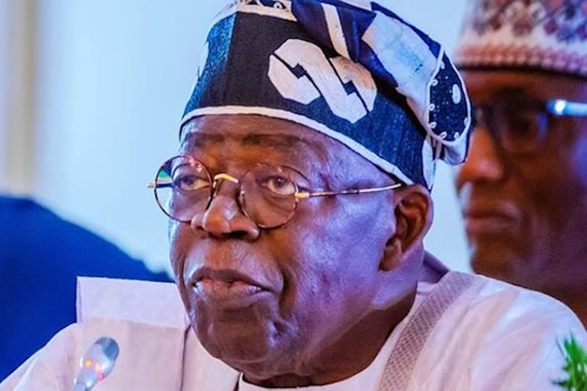 Elder man wearing a patterned head wrap and glasses, speaking into a microphone at an event.