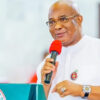 Man in white traditional attire speaks into a red microphone at a podium with a government seal visible on the front edge.