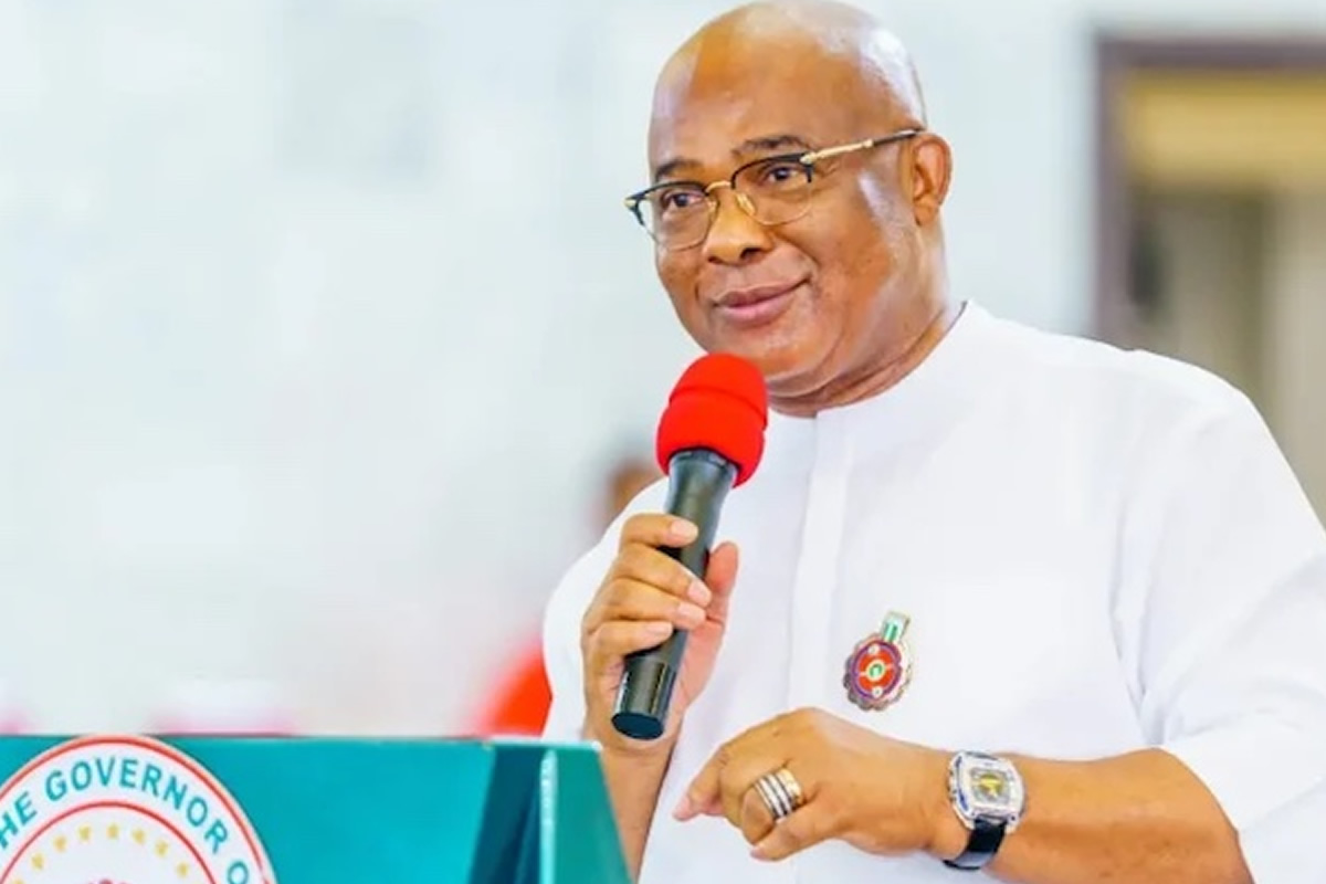 Man in white traditional attire speaks into a red microphone at a podium with a government seal visible on the front edge.