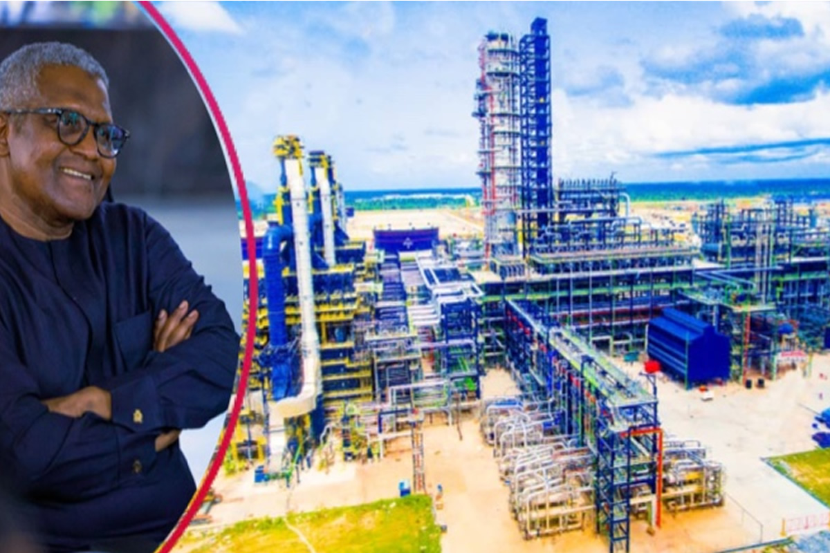 Smiling man in a dark blue shirt stands beside a large oil refinery complex by the seaside.